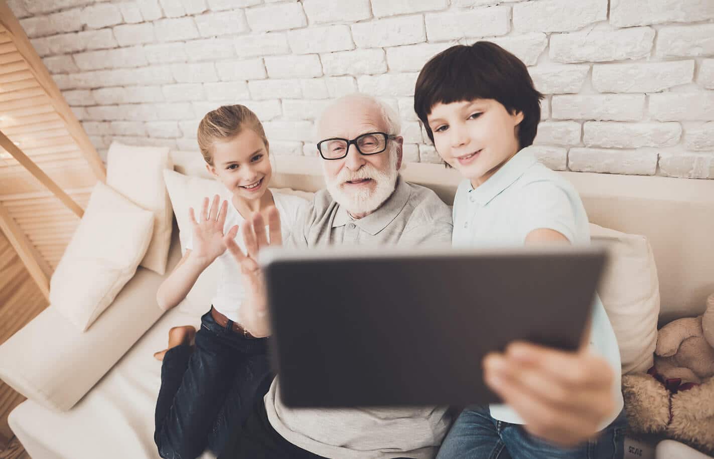 Senior man wearing black glasses sitting on a couch with a young boy and girl waving at a tablet