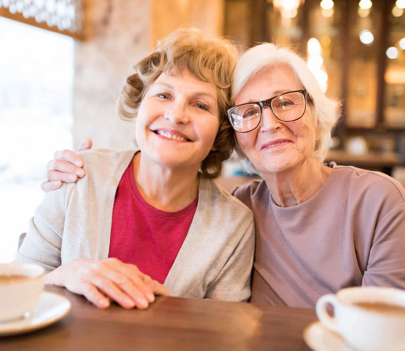 Senior mother and daughter with arms around each other, smiling at a coffee shop