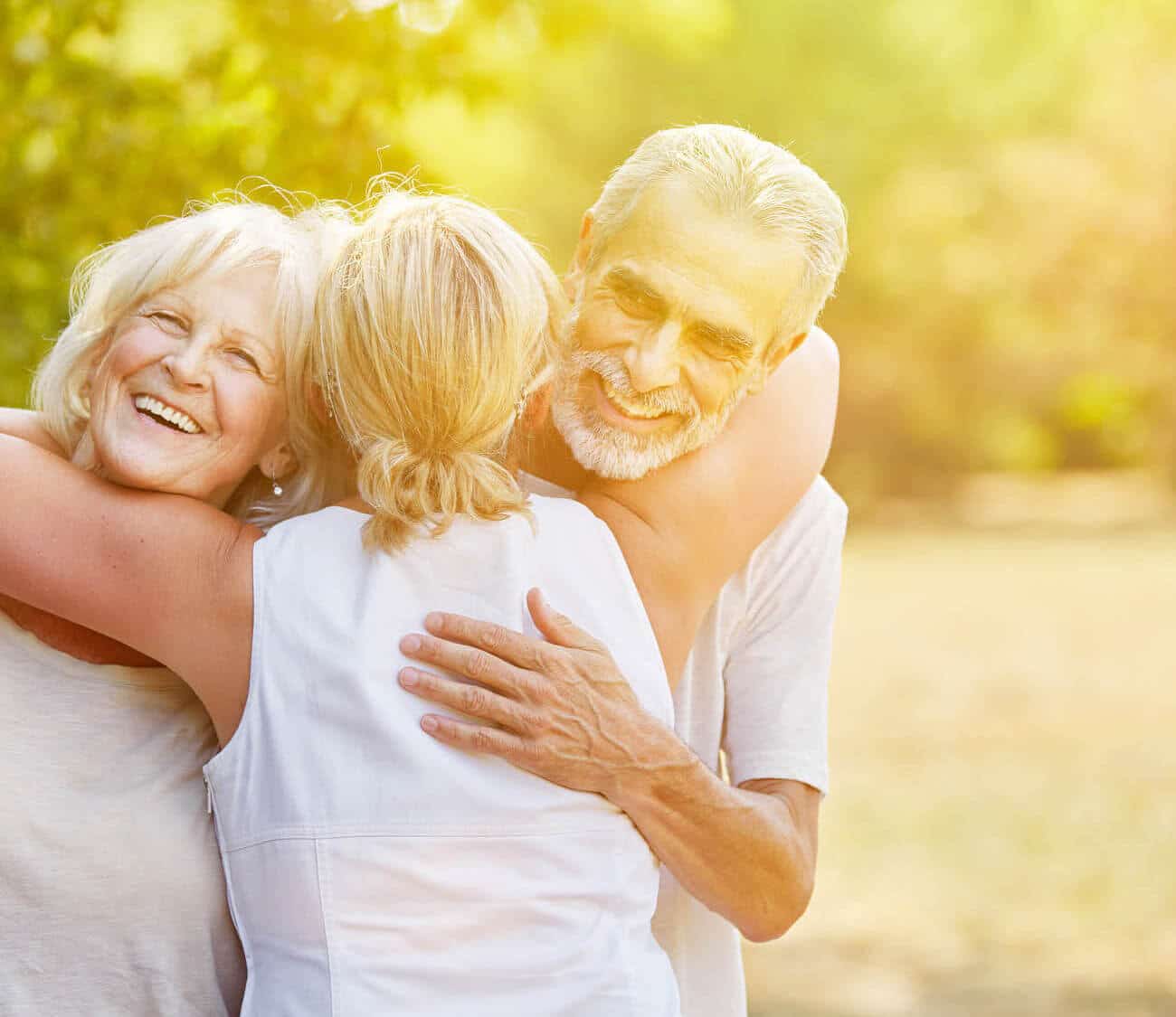 Adult daughter hugging both her senior parents at the same time in an orchard of trees.