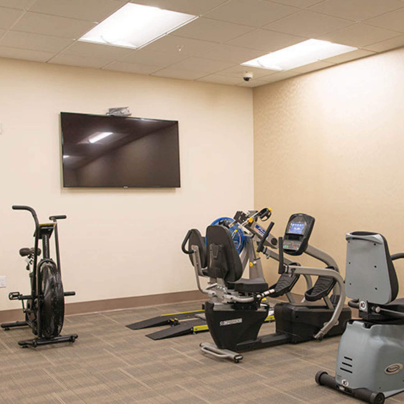 Workout room with large TV and multiple fitness machines, including treadmill, stationary bike among others.