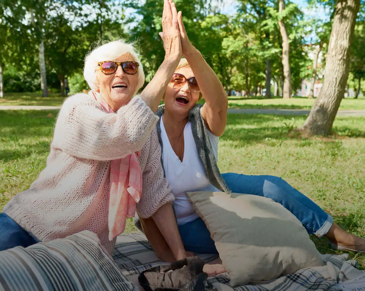 2 elderly women sitting on a blanket in the park high-fiving